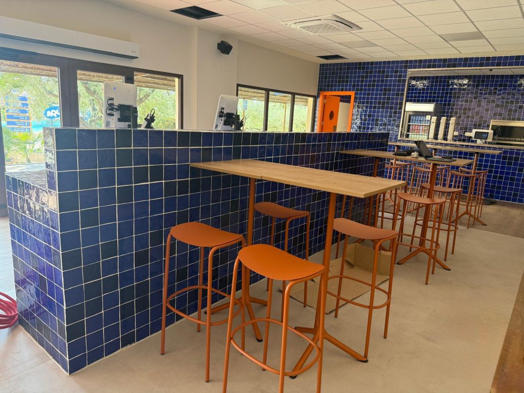 Cafe interior with blue tiled walls, tall wooden tables, and orange bar stools by a tiled half-wall near windows.
