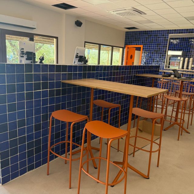 Cafe interior with blue tiled walls, tall wooden tables, and orange bar stools by a tiled half-wall near windows.