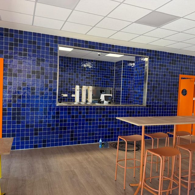 Cafeteria area with blue tiled walls, orange doors, and brown-topped tables with orange stools