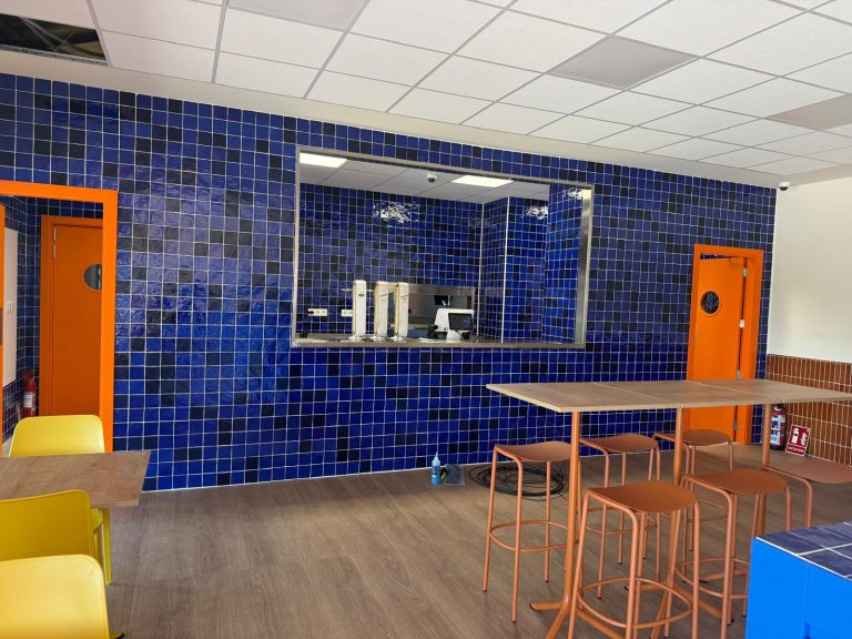 Cafeteria area with blue tiled walls, orange doors, and brown-topped tables with orange stools