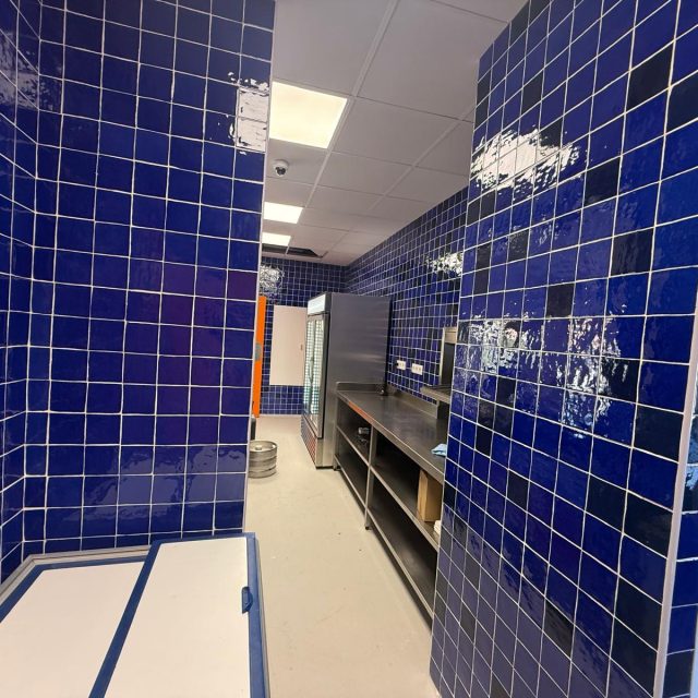 Narrow commercial kitchen with glossy blue tiled walls, stainless steel prep counter, and a glass-door refrigerator in the back.