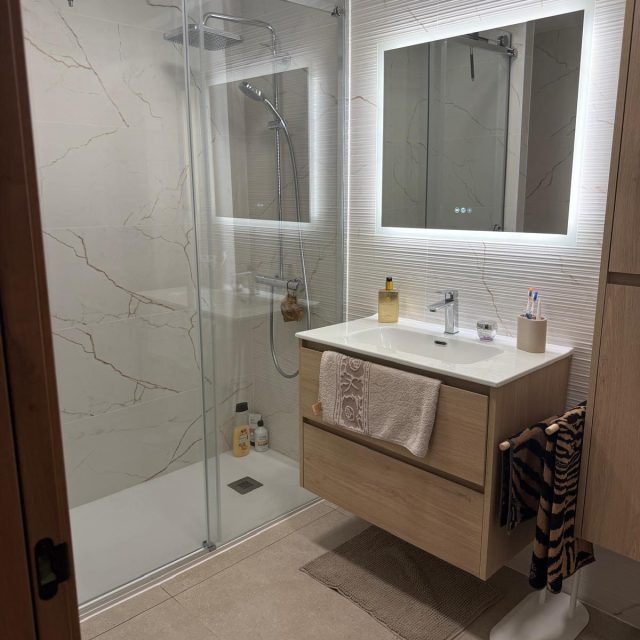 Modern bathroom with glass shower enclosure, marble walls and a wooden vanity beneath a backlit mirror.