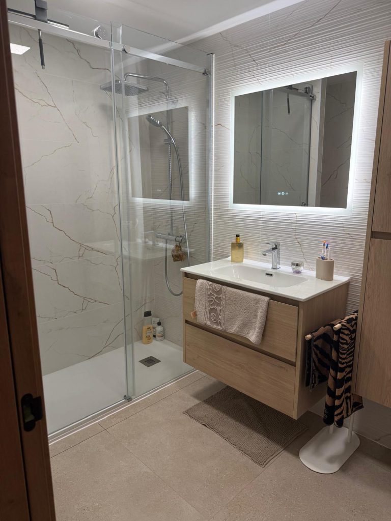 Modern bathroom with glass shower enclosure, marble walls and a wooden vanity beneath a backlit mirror.